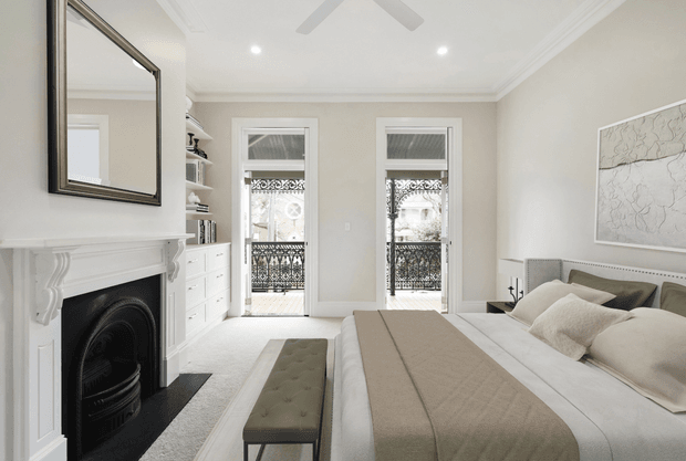 Bright, refined bedroom with a large upholstered bed, neutral bedding, an olive upholstered bench, built-in white shelving and drawers, a decorative white fireplace, tall French doors with ornate balcony railings, and a large wall mirror. The room has high decorative ceilings, recessed lighting, and a calm neutral palette.