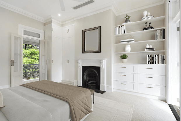 Bright, refined bedroom with a neutral upholstered bed, quilted taupe throw, white bespoke cabinetry, open display shelving, a decorative fireplace with traditional mantel, and a framed mirror. French doors open to a balcony with greenery beyond. The room has high-quality built-in joinery, soft carpet, and abundant natural light.