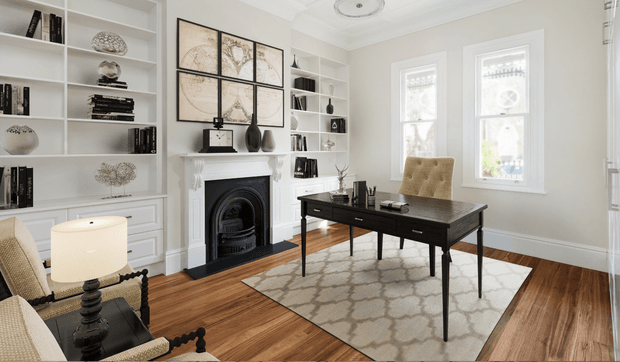 Elegant traditional home office with a dark timber desk and upholstered chair positioned on a patterned area rug. The room features warm timber flooring, white built-in shelving and cabinetry, a white decorative fireplace surround with black insert, tall sash windows, detailed ceiling mouldings, and carefully styled books and accessories.