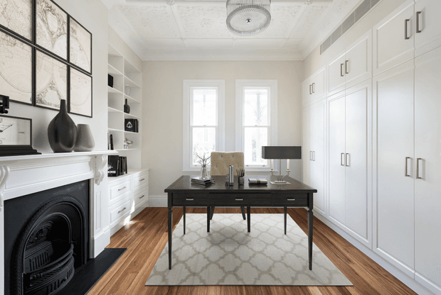 Bright, refined home office with a traditional fireplace, extensive white built-in cabinetry, a dark wood and black desk, patterned area rug, timber flooring, tall windows, and an ornate decorative ceiling with a central pendant light. The room combines classic architectural detailing with restrained contemporary furnishings.