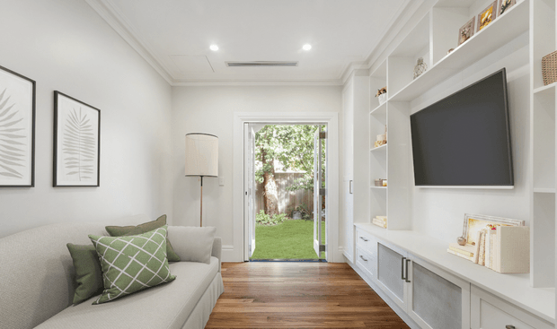 Bright, refined living room with a light upholstered sofa, green patterned cushions, framed botanical artwork, a floor lamp, warm timber flooring, and custom white built-in shelving and cabinetry incorporating a wall-mounted television. The room opens through glazed double doors to a small landscaped garden, with recessed lighting and decorative ceiling detailing.