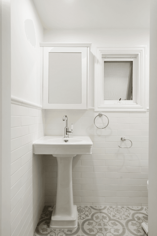 A compact, bright white bathroom with a traditional pedestal basin, chrome tapware and towel rings, white subway-tile wall lining, ornate patterned floor tiles, decorative white-framed mirror cabinetry, and a small frosted window with layered trim.