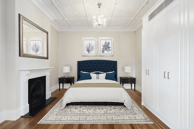 Elegant traditional bedroom with a centered upholstered bed, dark navy tufted headboard, white bedding, bedside tables and lamps, a decorative fireplace, built-in white wardrobes, framed botanical artwork, a patterned rug, timber flooring, and an ornate pressed ceiling with chandelier.