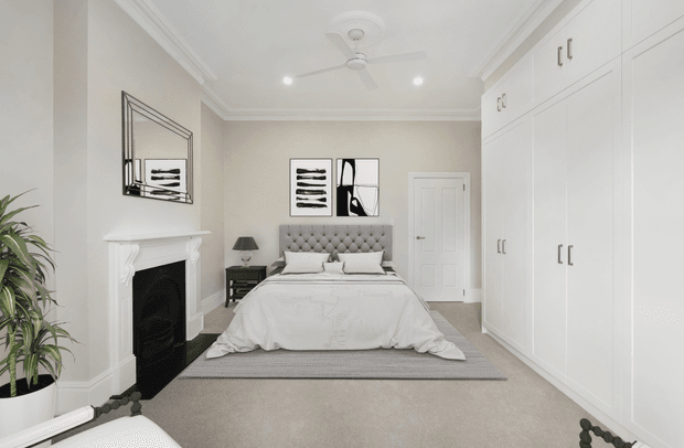A bright, elegant bedroom with a large upholstered tufted bed centred against the rear wall, extensive full-height white built-in wardrobes along the right side, a decorative white fireplace on the left, and neutral carpeting. Classic moulded cornices, ceiling medallion, recessed lighting, and a ceiling fan create a refined traditional interior, complemented by monochrome artwork and soft grey-beige furnishings.