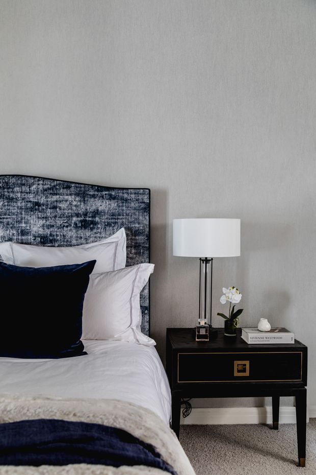A close-up view of a refined bedroom featuring a bed with white bedding, a deep navy velvet accent pillow and throw, and a textured blue-grey upholstered headboard. A black bedside table with brass detailing supports a modern white drum lamp, a small orchid, decorative objects, and books. The room has a softly textured light grey wall, pale carpet, and a restrained navy, white, grey, black, and brass palette.