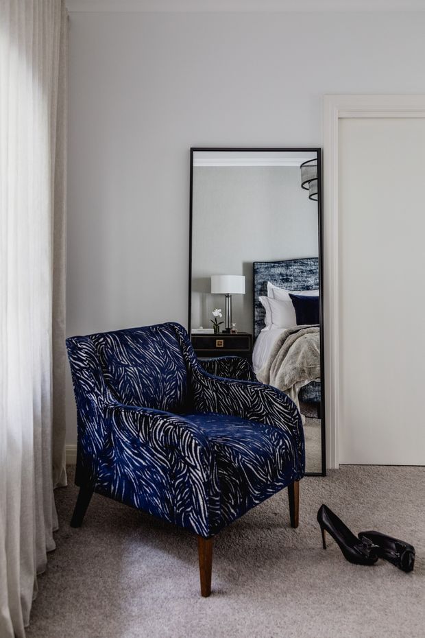 A refined bedroom corner with a navy patterned upholstered armchair positioned beside a full-height black-framed mirror. The mirror reflects a bed with a textured blue headboard, white bedding, a dark bedside table, and a table lamp. The room has pale grey walls, soft grey carpet, sheer curtains, and a white door, creating a calm, elegant interior.