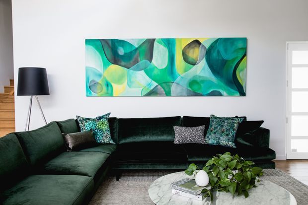 A contemporary living room with a deep green velvet L-shaped sectional and matching dark sofa, patterned accent cushions, a black tripod floor lamp, a large abstract turquoise and green artwork, a round marble coffee table, and leafy indoor plants. The room has pale walls, timber flooring, and a modern glazed entry door.
