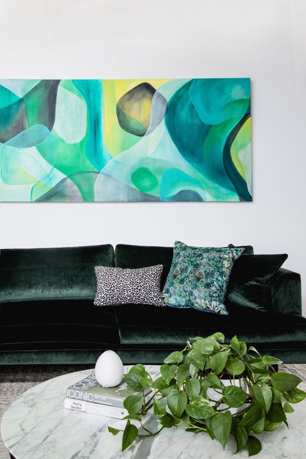 A styled living room vignette featuring a deep green velvet sofa with patterned decorative cushions, a round white marble coffee table, a leafy trailing houseplant, and a large abstract artwork in turquoise, green, yellow, and black tones on a white wall. The space has a bold, refined, eclectic feel.