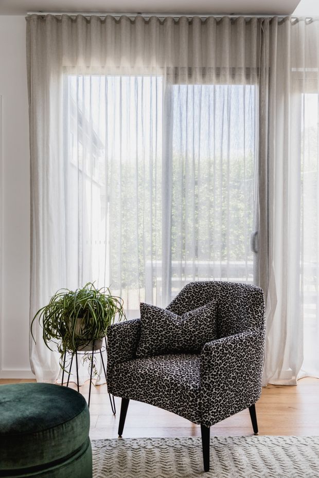 A bright, intimate living-room corner featuring a leopard-print upholstered armchair with matching cushions, a dark green round ottoman, and a potted plant on a slender black stand. Sheer white curtains span large glazing, allowing abundant daylight into the room. Pale timber flooring and a textured neutral rug complete the setting.