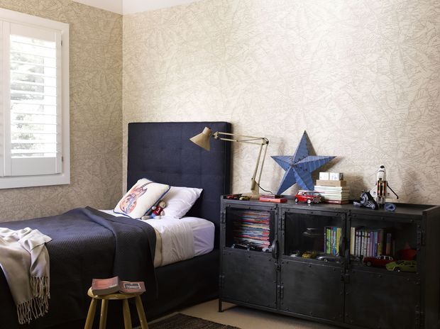 A cozy child's bedroom with a single bed, tall navy upholstered headboard, dark bedding, patterned beige wallpaper, white plantation shutters, and a black industrial-style storage cabinet filled with books and toys. A brass desk lamp, blue star decoration, and small stool add character.