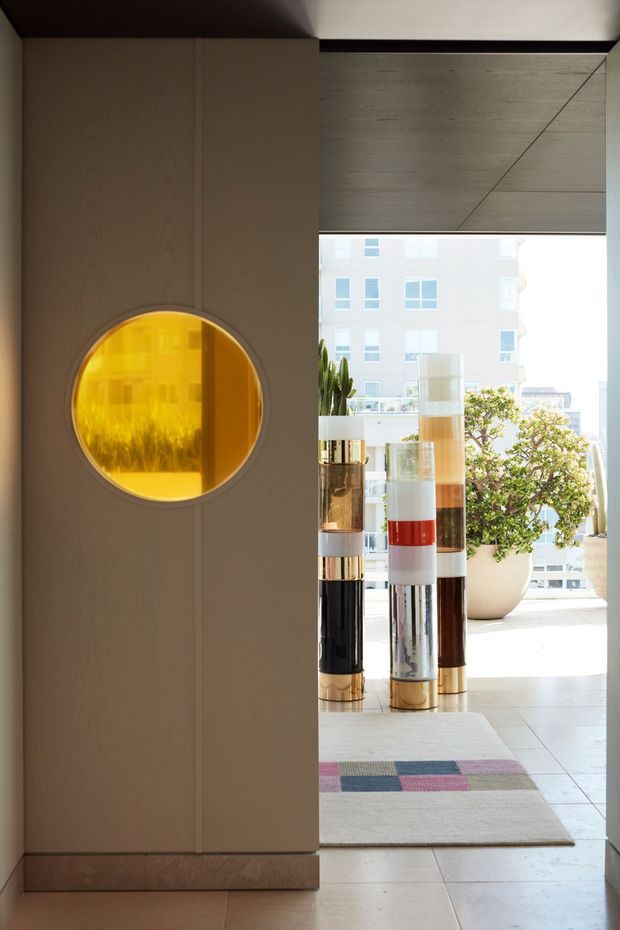 A refined apartment entry or hallway featuring a tall wood-paneled partition with a circular amber-glass inset. Beyond it is a bright glazed living area with sculptural cylindrical decorative screens, a large potted plant, pale tiled flooring, and a muted geometric rug. The space has strong natural light, warm timber tones, and polished contemporary detailing.
