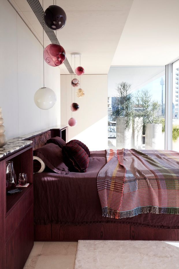 A refined bedroom with a built-in dark timber bed platform and matching side cabinetry, upholstered in deep plum bedding and layered with a multicoloured checked throw. A dramatic row of suspended, translucent spherical pendant lights hangs beside the bed. The room has pale walls and ceiling, a patterned stone-look benchtop or ledge, large glazed openings with city views, and a soft neutral rug over pale tiled flooring. The overall composition is warm, eclectic, and luxurious with strong sculptural lighting.