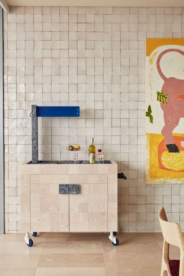 A compact, characterful dining or entertaining area featuring a pale timber mobile drinks cabinet with blue caster wheels, integrated dark stone or granite benchtop and a sculptural blue wall-mounted tap or shelf element. The entire wall is clad in small, irregularly aligned pale ceramic tiles with visible grout lines. A large colourful abstract artwork hangs at right, with part of a timber dining chair visible in the foreground. The palette is warm and playful, combining pale wood, cream tile, blue accents and vivid artwork.
