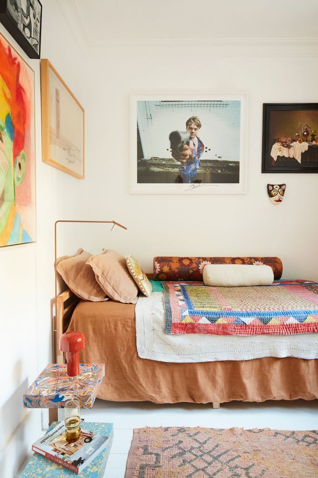 A warmly styled bedroom with a bed dressed in layered terracotta-brown linens, patterned quilt, assorted cushions, and a vintage upholstered headboard. White walls with ornate ceiling moulding are covered in colourful framed artwork, while a small sculptural wall mask, slender floor lamp, red table lamp, patterned side table, books, and a geometric rug add eclectic character. The room is bright and intimate with a richly layered, collected aesthetic.