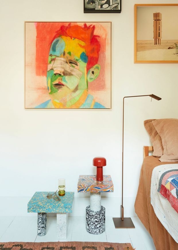 A warmly styled bedroom corner with a low bed at right, terracotta bedding and a colorful patchwork quilt. The pale wall displays several framed artworks, including a large vibrant abstract portrait. Two small sculptural side tables with patterned tops, a red table lamp, and a slender dark floor lamp create an eclectic, artistic arrangement. The room has a light-colored hard floor and a partial patterned rug.