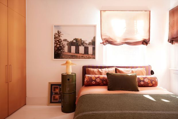 Warm, compact bedroom with a built-in wood wardrobe, low bed, layered patterned pillows, and a textured brown bedcover. Soft daylight enters through a fabric Roman blind, while a framed photograph and small cylindrical side table add restrained decorative detail.