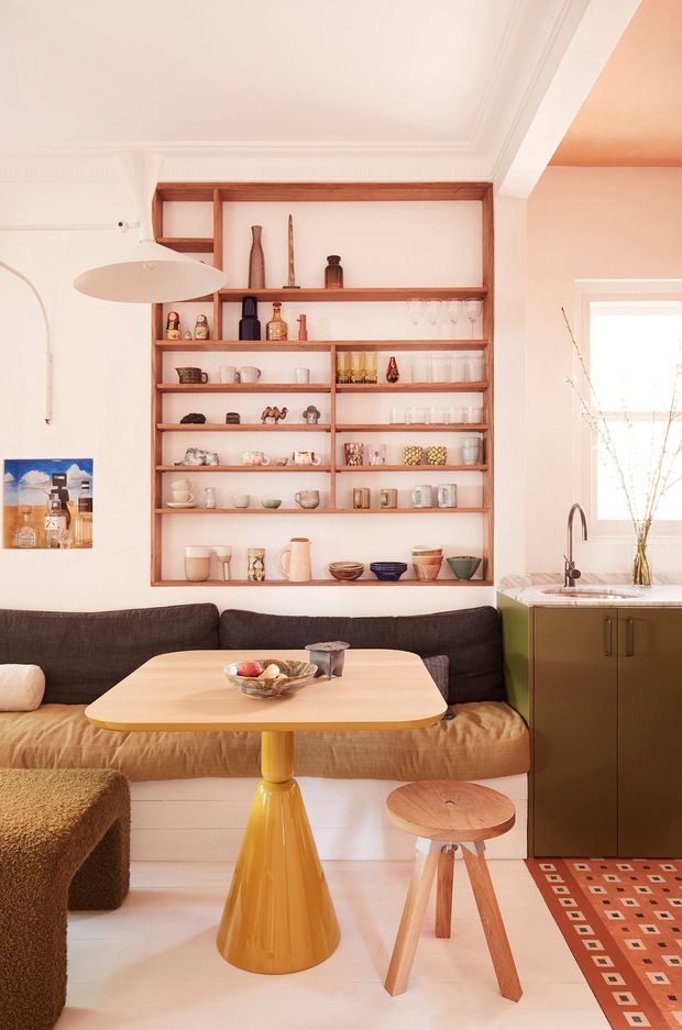 A warm, compact open-plan dining and living area with a built-in timber display shelf filled with glassware and ceramics. A dark upholstered banquette wraps around a light timber dining table with a distinctive yellow pedestal base, accompanied by a small wooden stool. To the right is a compact green kitchen cabinet with a stone benchtop and a curved metal tap. The room features white walls and ceiling with decorative cornice, pale flooring, warm timber shelving, and colourful patterned textiles.
