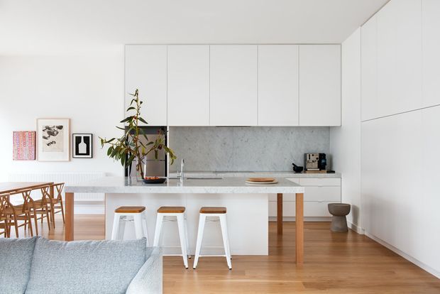 Bright open-plan contemporary kitchen and dining area viewed from the living room. The space features streamlined white flat-panel cabinetry, a pale stone benchtop and backsplash, timber flooring, a central island with three stools, a timber dining table and chairs, and restrained framed artwork. A compact living-room sofa is visible in the foreground.