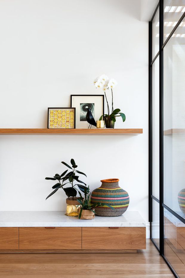 A bright minimalist interior vignette featuring a floating timber display shelf with framed artwork, a black bird sculpture and orchid, above a low built-in timber cabinet with a pale stone top. A colourful patterned ceramic vessel and greenery sit on the cabinet. Full-height black-framed glazing or a glazed internal partition runs along the right side, reflecting the cabinetry and timber floor.