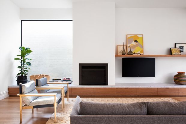 Bright contemporary living room with a white plaster fireplace wall, built-in timber media cabinetry, wall-mounted television, floating display shelf, large glazed opening, timber flooring, and a mix of neutral upholstered seating and warm wood accents.