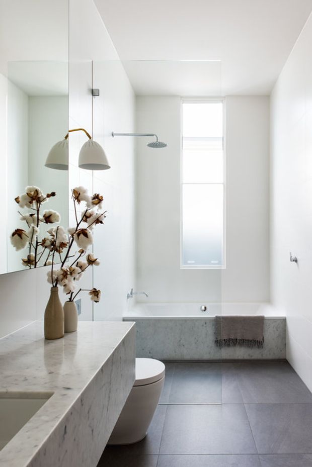 Bright, minimalist contemporary bathroom with a freestanding-style built-in bathtub, wall-mounted shower, large frameless mirror, and a marble vanity with integrated basin. White walls and ceiling contrast with grey stone-look floor tiles, while a narrow window provides natural light. A small vase of cotton branches adds a soft organic accent.