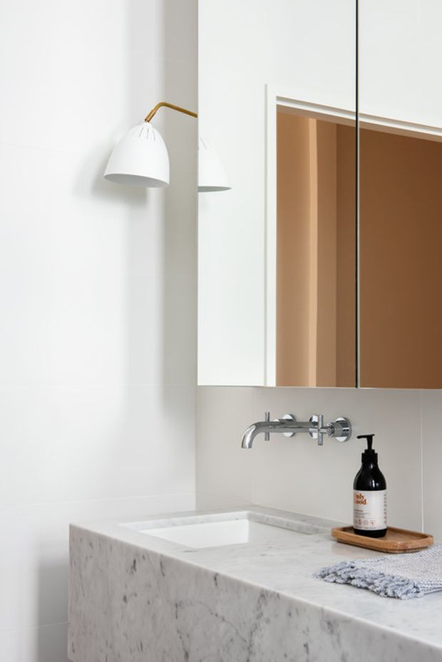 A bright, minimalist bathroom vanity with a thick white marble countertop and integrated rectangular basin. A wall-mounted chrome faucet sits beneath a mirrored cabinet, while a white wall lamp with a brass arm and a few restrained countertop accessories add detail. The reflected warm-toned doorway or cabinetry provides subtle contrast.
