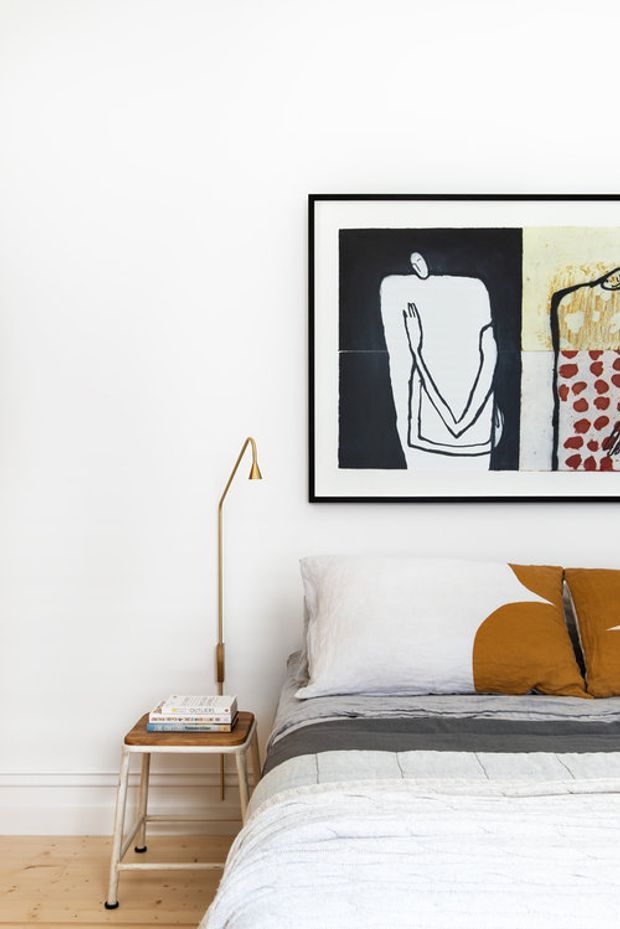 Bright, minimalist bedroom vignette with a partially visible bed dressed in white and grey bedding, mustard geometric cushions, a slim brass-toned floor lamp, a small timber bedside table with books, pale timber flooring, and bold framed abstract artwork above the bed.