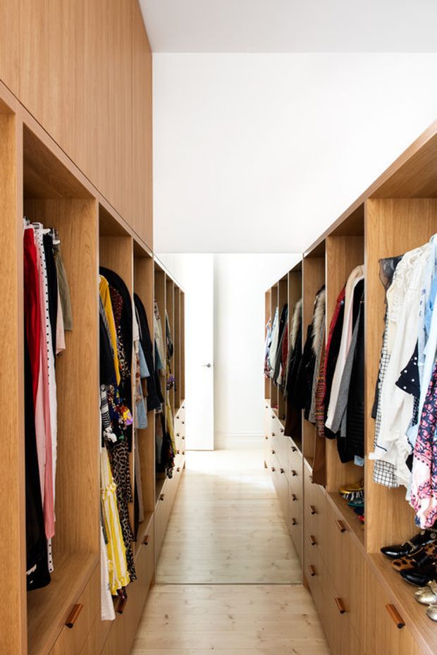 Bright walk-in wardrobe with symmetrical built-in timber cabinetry lining both sides of a narrow passage. Open hanging rails display clothing, with drawers and shelving below, pale timber flooring, a white ceiling, and a full-height mirror or glazed opening at the far end.