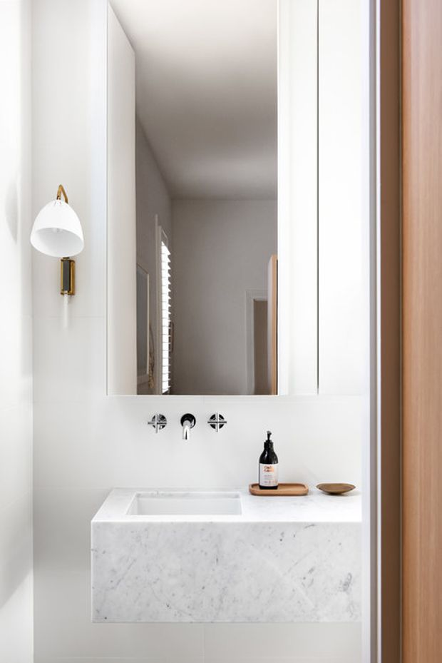 A compact, bright bathroom vanity with a wall-mounted white marble or stone basin and benchtop, polished chrome wall-mounted tapware, a large frameless mirror, and a white dome wall sconce with brass detailing. White tiled walls and a warm timber door frame create a clean, refined look.