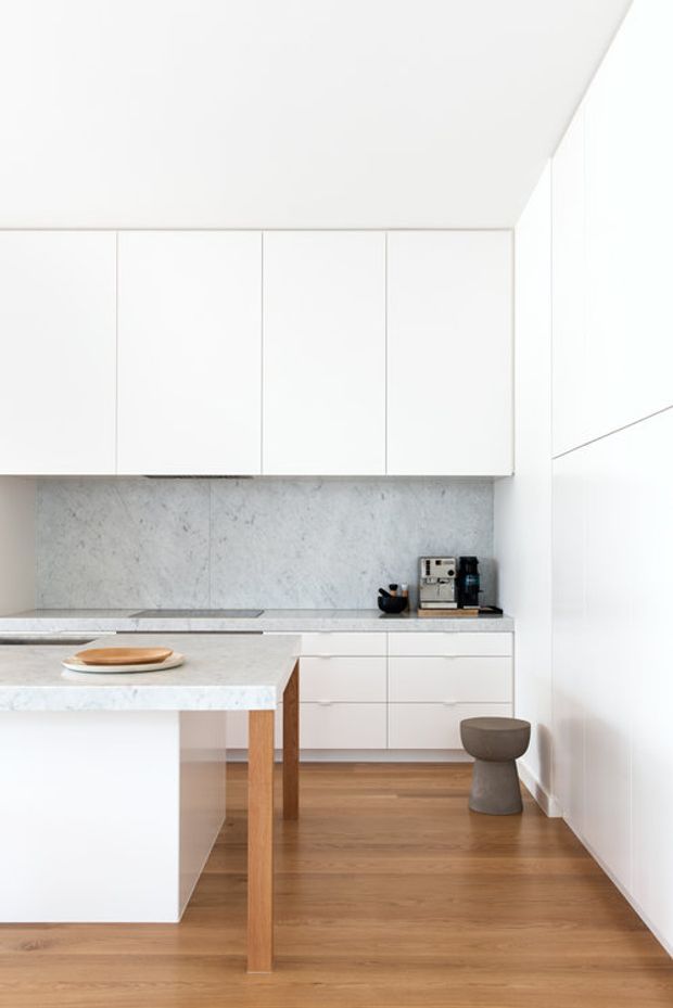 A bright, minimalist contemporary kitchen with seamless white full-height cabinetry, a pale grey veined stone splashback and benchtops, integrated appliances, and a compact island with timber supports. Warm timber flooring contrasts with the white joinery, while a sculptural grey stool and coffee machine provide restrained accents.