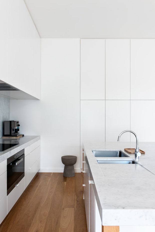 A bright, minimalist galley kitchen with full-height white cabinetry, a large pale stone island with an integrated double sink and brushed metal tapware, and built-in black appliances along the opposite wall. Warm timber flooring contrasts with the predominantly white, clean-lined fit-out. A small dark sculptural stool sits at the end of the kitchen.