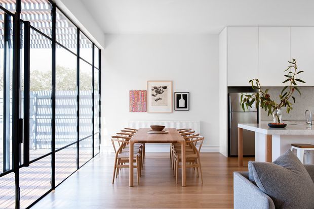 Bright open-plan dining and kitchen interior with a large timber dining table and matching wooden chairs, light timber flooring, white walls and cabinetry, and expansive black-framed glazed doors along one side. A grey sofa is partially visible in the foreground, with restrained artwork and a potted branch adding subtle decoration.