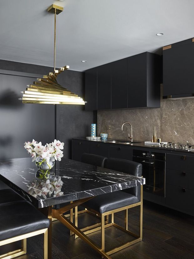 A sophisticated dark-toned kitchen and dining area with matte black cabinetry, a dramatic veined stone backsplash and island benchtop, dark timber flooring, and a sculptural brass-and-glass pendant light. Black upholstered dining chairs with brass frames surround the island, while a polished floral arrangement adds a contrasting decorative accent.
