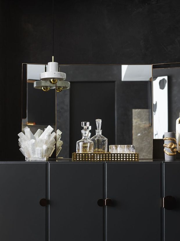 A dark, moody interior vignette featuring a black built-in sideboard or bar cabinet with round pulls, framed mirror panels, crystal decanters and tumblers on a brass-studded tray, a sculptural white pendant light, and decorative crystal objects. The styling is sophisticated and highly contrasted, with black surfaces, reflective glass, and warm metallic accents.