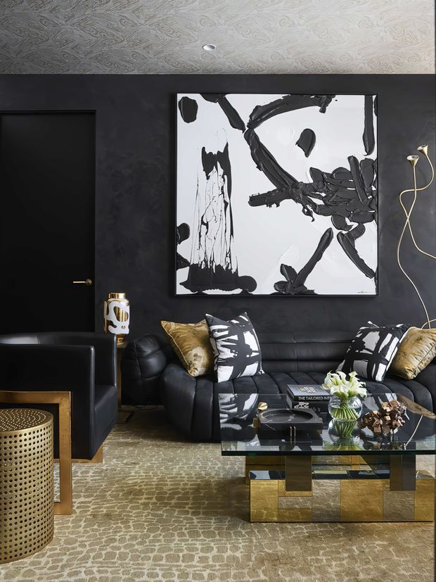 A sophisticated dark-toned living room featuring a black leather sofa and armchair, patterned black-and-white cushions, metallic gold accent pillows, and a glass coffee table with a geometric gold-and-black base. The room has charcoal textured walls, a black door, a sculptural black-and-white abstract artwork, a brass wall light, a patterned carpet, and a decorative gold-toned perforated side table. The overall aesthetic is dramatic, luxurious, and art-focused.