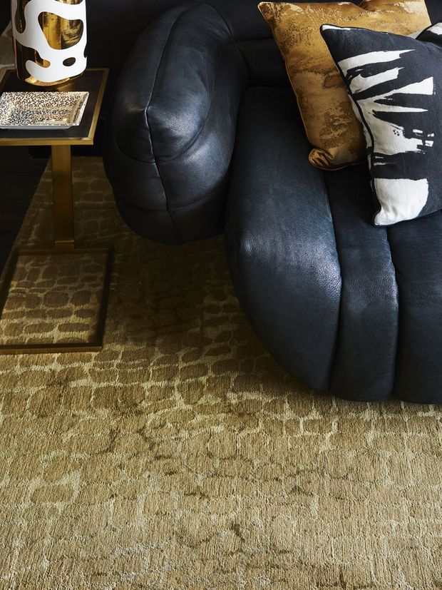 Close-up interior detail of a sophisticated living room, featuring a large black leather sofa with rounded upholstered forms, patterned gold and black-and-white cushions, a textured tan carpet, and a small dark side table with a decorative lamp and book. The composition emphasizes rich textures, dark tones, and layered patterns.