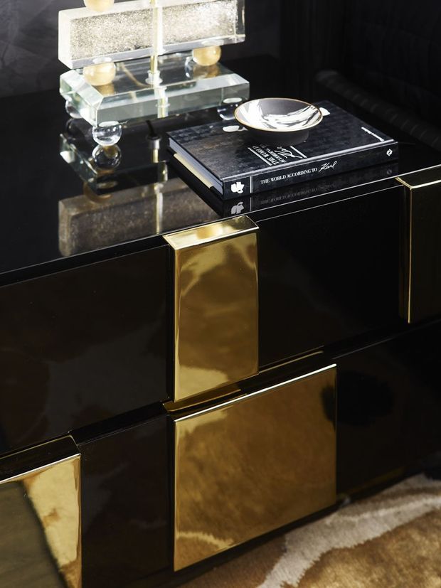 Close-up interior detail of a highly polished black sideboard or console with geometric brass-gold inset panels and trim. A glass-and-metal decorative lamp, books, and a small dish sit on the reflective top, creating a luxurious, high-contrast composition.
