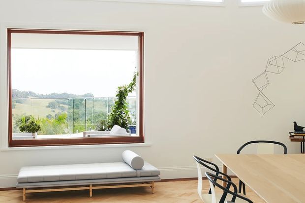 Bright, minimalist dining and living area with a large timber-framed picture window overlooking a green landscape. The room features a pale timber dining table with black metal chairs, a simple upholstered daybed, light timber flooring, white walls, indoor plants, and understated geometric wall art.