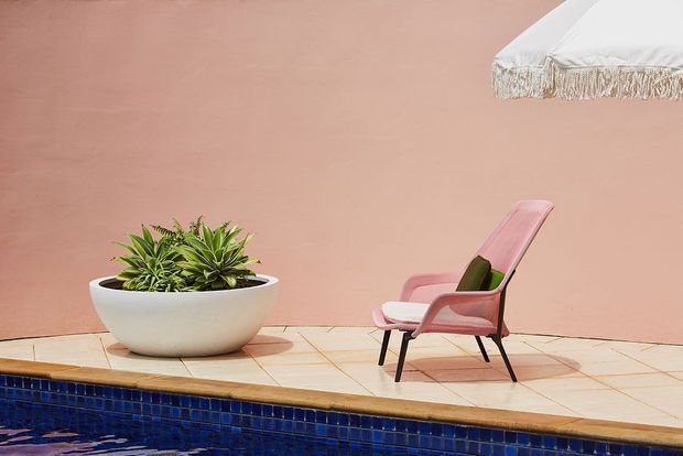 A sunlit poolside terrace with a dusty pink rendered wall, a large white planter containing succulents, and a pink reclining lounge chair beneath the edge of a fringed white parasol. Pale tiled paving surrounds the deep blue tiled swimming pool.