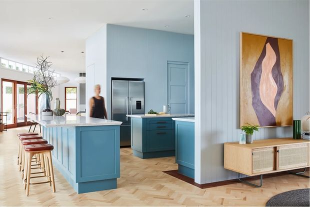 Bright open-plan kitchen and living area with blue painted cabinetry, a central island with bar stools, stainless-steel appliances, pale blue vertical wall panelling, and a warm herringbone timber floor. A timber-framed glazed door opens toward an exterior area, while a sideboard and abstract artwork occupy the adjacent living space.