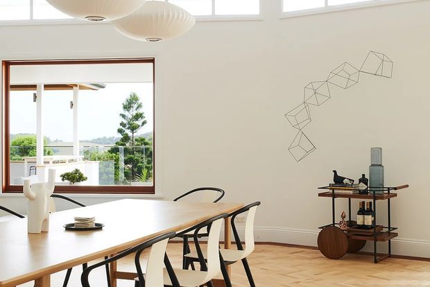 Bright contemporary dining room with a long light-wood dining table and sculptural white and black chairs. The room has white walls, high clerestory windows, a large timber-framed picture window opening to greenery, pale timber parquet flooring, sculptural pendant lights, and a compact open drinks bar with decorative objects and bottles. A geometric wire wall sculpture adds a subtle feature.