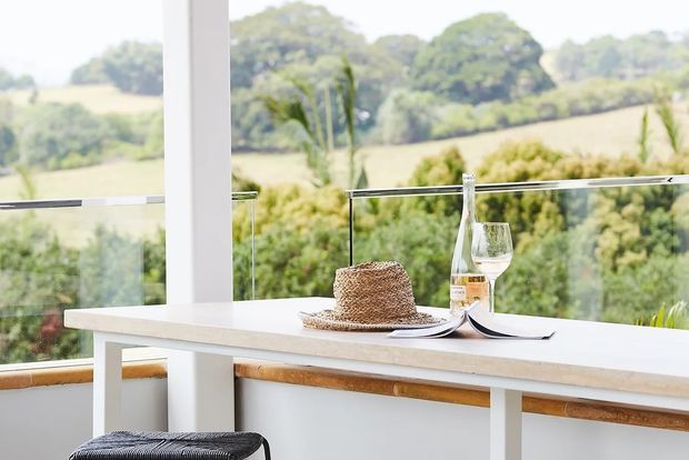 Bright, airy interior with a slim white table or built-in counter positioned beside floor-to-ceiling glazing and a frameless glass balustrade. A woven hat, open magazine, bottle and wine glass sit on the surface, while lush landscaped countryside is visible beyond the windows. The space has a clean contemporary feel with white structural elements and warm timber trim.