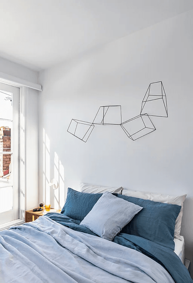 Bright, compact bedroom with a neatly made bed dressed in layered pale blue and deep blue bedding, white pillows, and a simple wooden bedside table. White walls and ceiling create an airy setting, with strong daylight entering through a tall glazed door or window. A geometric wire-style wall sculpture decorates the wall above the bed.