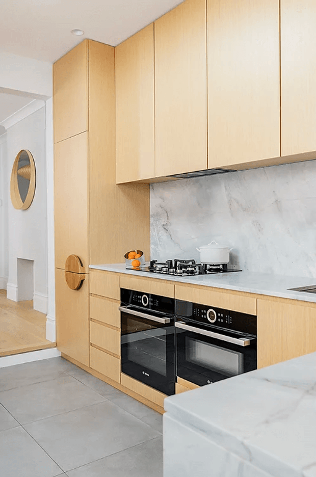Bright contemporary kitchen with extensive light timber cabinetry, integrated black double ovens, a gas cooktop, and a pale veined stone backsplash and island benchtop. Large grey floor tiles and a round timber-framed opening add detail, while the restrained palette creates a clean, refined atmosphere.