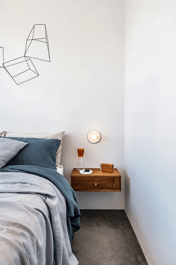 A compact, minimalist bedroom corner with a partially visible bed dressed in pale blue-grey and deep blue bedding, a wall-mounted dark timber bedside drawer, a small round wall light, and geometric black line artwork on a white wall. Dark carpet and clean, uncluttered detailing create a calm contemporary feel.