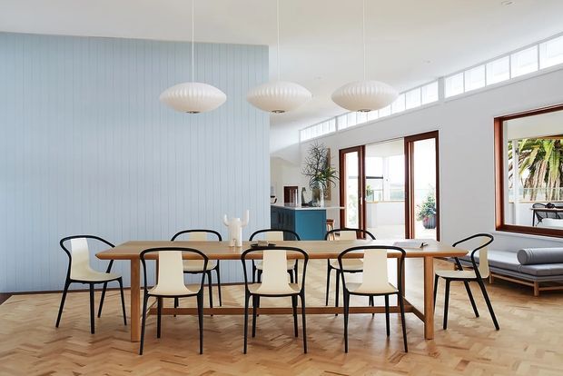 Bright, airy open-plan dining area with a long timber dining table and mixed black-framed, cream upholstered chairs. The room features a pale blue vertical-panel feature wall, warm parquet timber flooring, large timber-framed glazed doors and windows, and three sculptural pendant lights. A compact blue kitchen or joinery area is visible beyond the dining space, with a lounge area seen through the right-hand opening.