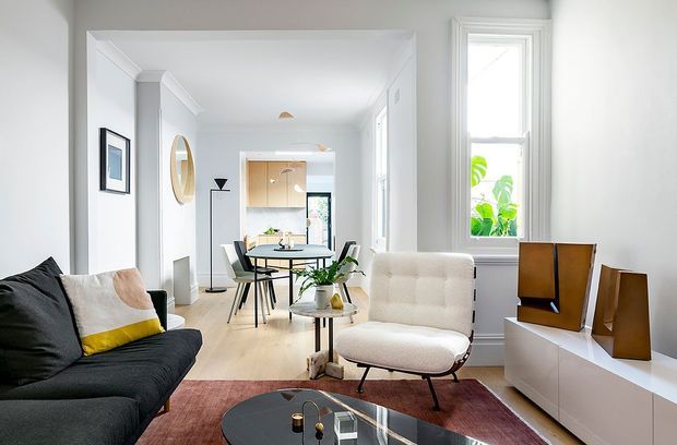Bright, refined open-plan living and dining area with white walls, tall windows, a dark sofa, sculptural upholstered lounge chair, round dining table, and low white media cabinetry. A compact kitchen is visible at the rear, with warm timber cabinetry and indoor plants adding contrast.