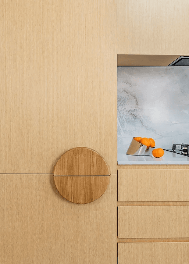 Close-up view of a minimalist timber-lined kitchen or joinery wall, featuring a round flush-mounted timber door or handle detail, integrated timber drawers, and a small opening revealing a pale stone benchtop and splashback with oranges and a cooktop beyond.