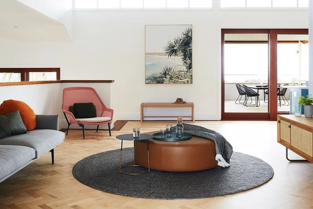 Bright, airy mid-century-inspired living room with a curved grey sofa, sculptural lounge chair, round leather ottoman coffee table, dark circular rug, timber parquet flooring, and warm timber-framed glazing. A dining or outdoor seating area is visible beyond the glazed doors, with high clerestory windows bringing in abundant natural light.