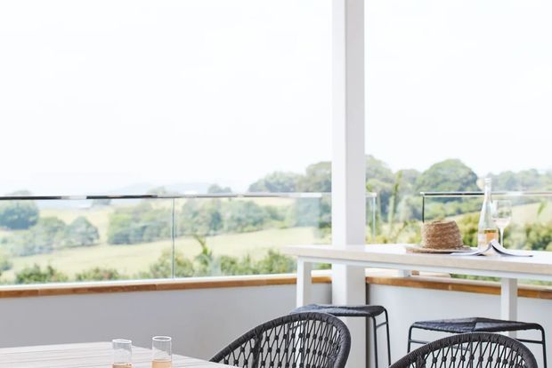 Bright outdoor terrace or balcony with a glass balustrade, white structural posts and a white bar-height table overlooking a green rural landscape. Two woven dark chairs and simple table settings create a relaxed dining or entertaining area.